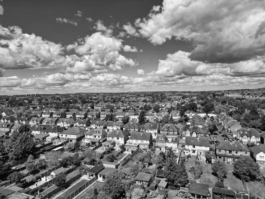 A black and white aerial photograph of a suburban neighbourhood under a partly cloudy sky, with rows of detached and semi-detached houses, each with small gardens and fenced yards, stretching across t