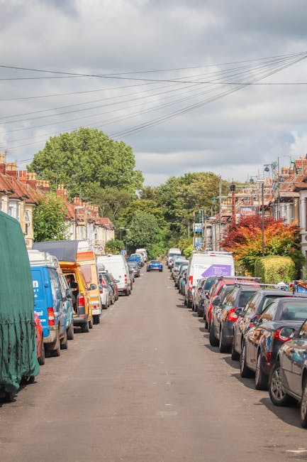 A residential street scene during daytime showing rows of parked cars lining both sides of the road, with a variety of small vans and moving trucks parked in the available space. The vehicles include a blue van covered with a green protective tarp and several white, orange, and purple moving vans, some with open rear doors revealing loaded packing boxes. The street is flanked by terraced houses with brick facades, small gardens, and trees, some showing foliage changes indicative of spring or summer. Overhead, power lines stretch across a cloudy sky, and the street appears to be a typical urban or suburban area used for home relocation and furniture transport, with Man with Van Tulse Hill potentially providing such services related to this scene, which involves the logistics of loading or unloading household items during a house move or local removal process.