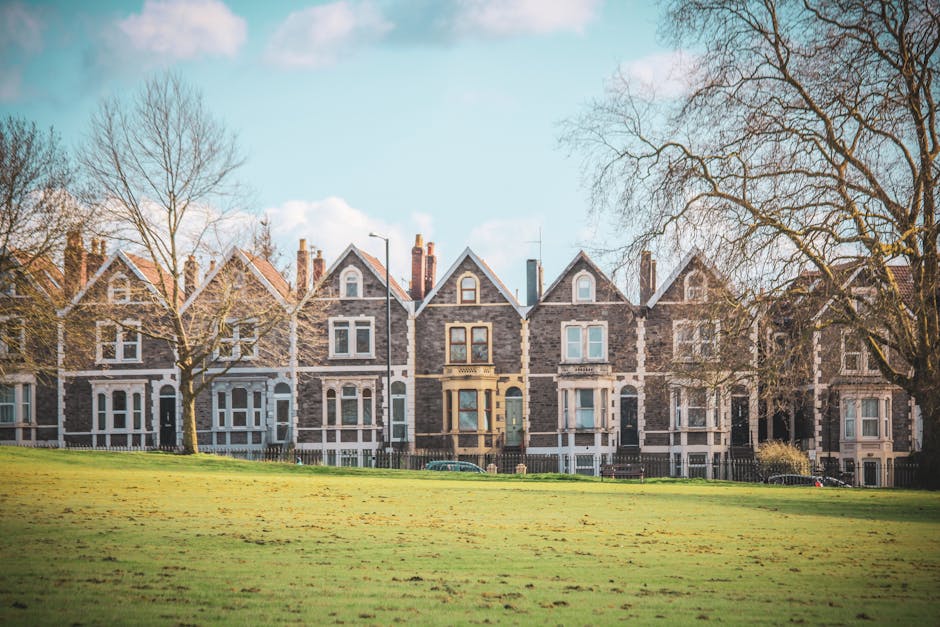 Image showing a row of traditional terraced houses with stone facades and pitched roofs, situated behind a well-maintained grassy area with a few bare trees indicating a late autumn or winter season. The houses are positioned along a street, with some cars parked in front, and the scene is captured during daylight with clear blue skies and soft clouds. In the context of home relocation, this setting reflects a residential area where Man with Van Tulse Hill might facilitate house removals or furniture transport services, involving loading and unloading of belongings from properties similar to these. The image emphasizes the exterior environment, with no visible interior or moving equipment present, but highlights the typical architecture and outdoor environment encountered during house removals in the Tulse Hill area.