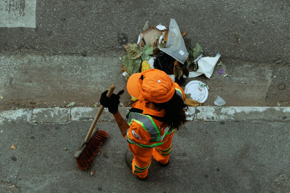 A woman wearing high-visibility orange workwear and an orange safety helmet is standing on a pavement, seen from above, working to clear a small pile of bulky waste including cardboard boxes, plastic packaging, paper, and other refuse against a concrete and brick wall. She holds a long-handled broom with a red brush, and her left hand gestures, possibly giving instructions or indicating the waste. The scene takes place near a doorway or entrance to a property, with the waste positioned on the sidewalk just outside. In the background, various waste materials such as crumpled paper, plastic bags, and packaging are visible, some partially covered by a plastic sheet. The lighting suggests outdoor daylight, suitable for tasks related to removals, household waste handling, or planning for a move. This image exemplifies the process of managing bulky waste, which could relate to house removals or dealing with waste as part of a home relocation service provided by Man with Van Tulse Hill.