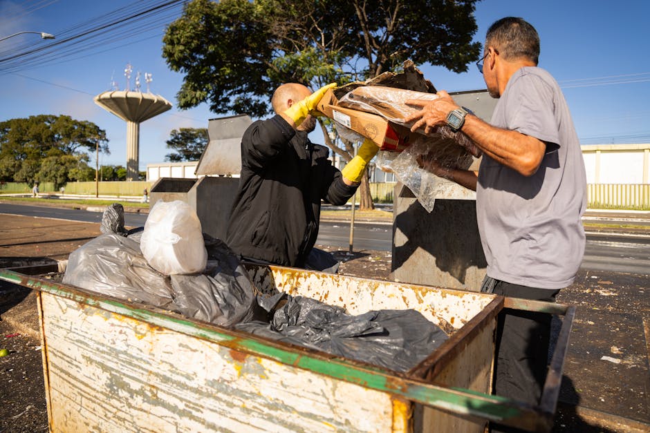 Two men are shown participating in the collection of bulky waste in an outdoor setting under clear blue skies. One man, wearing a black jacket and yellow gloves, is lifting a cardboard box filled with various items wrapped in plastic, while the other, dressed in a grey T-shirt and glasses, is assisting by holding the box steady. They are positioned near a large, rusty metal container filled with black and grey garbage bags, situated on a paved surface. The background features a large tree with green foliage, a water tower structure, and a white industrial or storage building with yellow fencing. This scene captures the process of loading and handling waste materials, relevant to home relocation or disposal services provided by companies like Man with Van Tulse Hill, as part of their removals and moving logistics services.