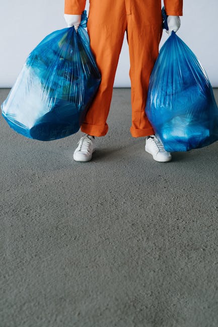 A woman wearing high-visibility orange workwear and an orange safety helmet is standing on a pavement, seen from above, working to clear a small pile of bulky waste including cardboard boxes, plastic packaging, paper, and other refuse against a concrete and brick wall. She holds a long-handled broom with a red brush, and her left hand gestures, possibly giving instructions or indicating the waste. The scene takes place near a doorway or entrance to a property, with the waste positioned on the sidewalk just outside. In the background, various waste materials such as crumpled paper, plastic bags, and packaging are visible, some partially covered by a plastic sheet. The lighting suggests outdoor daylight, suitable for tasks related to removals, household waste handling, or planning for a move. This image exemplifies the process of managing bulky waste, which could relate to house removals or dealing with waste as part of a home relocation service provided by Man with Van Tulse Hill.