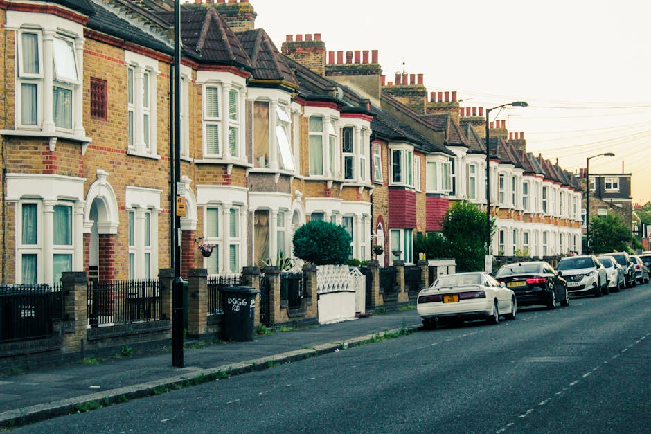 A row of terraced houses in Tulse Hill with brick facades and bay windows, some open with curtains visible inside. The houses are separated by small front gardens enclosed by low brick and metal fences, with a few with potted plants or small bushes. Parked cars line the street in front of the houses, and street lamps are mounted on tall poles along the pavement. The scene is captured during daylight with overcast sky, providing natural lighting. Visible in the foreground is a black wheelie bin labeled 'DOGG' situated beside a lamppost. This image relates to the process of home relocation and furniture transport, showing the environment where house removals by Man with Van Tulse Hill might take place, highlighting the typical street scene involved in local moves and the logistics of packing and loading objects for transport.