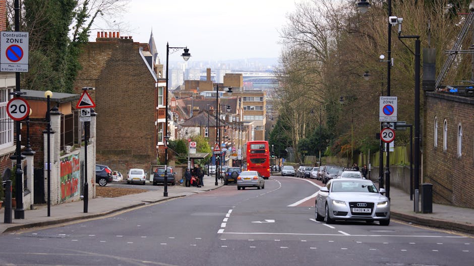 A busy residential street near Tulse Hill SW2 showing parked cars on both sides of the road and a red double-decker bus traveling away from the camera while moving downhill. In the foreground, a silver Audi vehicle is waiting at a pedestrian crossing with white dashed markings on the asphalt. To the left, there is a sidewalk with pedestrians, street signs indicating parking restrictions and a controlled zone, and a bus stop shelter. Further along the street, several cars are parked along the pavement, and additional street signs, lampposts, and trees line the sidewalks. The street is bordered by brick buildings and houses on either side, with some trees providing limited foliage, and the background reveals a view of the urban cityscape with various multi-storey buildings and infrastructure. Man with Van Tulse Hill may perform home relocation, furniture transport, packing, and loading services in this area, supporting efficient moving logistics.