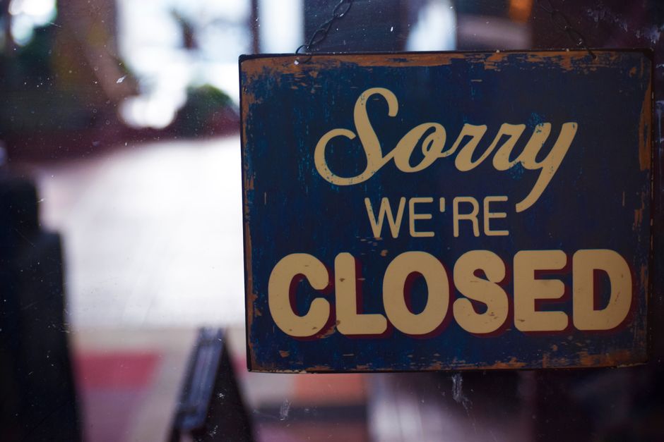 A rectangular blue sign with a distressed, weathered appearance hangs on a glass door or window. The sign displays the message, 'Sorry we're closed,' in large, beige-colored, cursive and block lettering. Behind the sign, there are blurred reflections and indistinct shapes, suggesting an indoor setting with some natural light coming from outside. This image captures the signage typically seen at a shop or office that may temporarily close due to parking suspensions or road closures near Tulse Hill SW2, relevant to home relocation or moving services. The sign's placement on glass indicates an entryway, which could be part of a moving company's premises like Man with Van Tulse Hill, supporting logistics and packing operations associated with house removals and furniture transport.