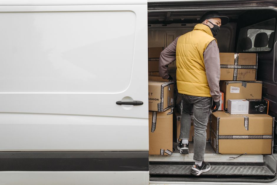 A man with a cap, white t-shirt, and jeans is walking alongside a white commercial van parked on a residential street, with the side door closed. The van is positioned parallel to the pavement in fron