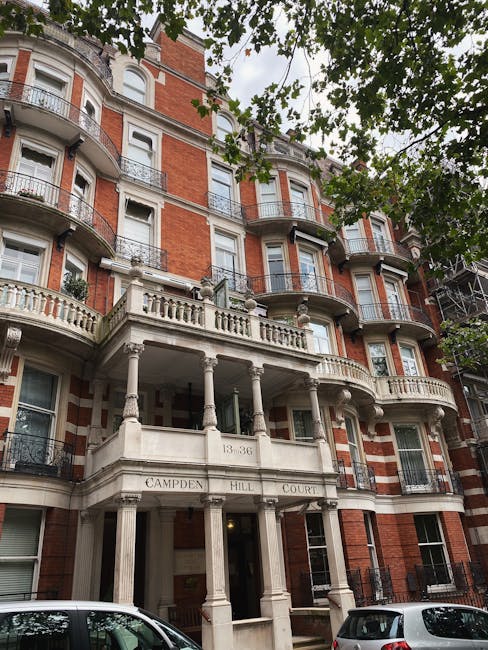 Outdoor view of a multi-storey residential building with a prominent white stone entrance featuring classical columns and decorative balustrades. The building's façade is constructed of red brick with white accents, and several balconies with wrought iron railings are visible on each floor. The upper sections are adorned with arched windows, and a tree with green leaves partially obscures the top of the building. Parked cars are visible in the foreground on the street pavement, indicating an active urban setting. This scene illustrates a typical example of a residential property in Tulse Hill, relevant to home relocations and furniture transport services. For house removals and packing logistics, companies like Man with Van Tulse Hill might manage the loading and unloading process, involving vehicles and equipment in such environments.