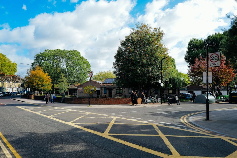 A street scene in Tulse Hill showing pedestrians crossing the road, some carrying bags, and a row of parked bicycles and scooters along the sidewalk. In the background, there are residential buildings with brick facades and dark tiled roofs, partially obscured by large green trees with autumn foliage. The street features yellow road markings and a speed limit sign indicating 20 mph. Street lamps and traffic signs are visible, and the overall scene suggests a busy urban area suitable for home relocation or moving services, with few vehicles and clear weather. Man with Van Tulse Hill may utilize such locations for loading or unloading furniture and boxes during a same-day move in Tulse Hill.
