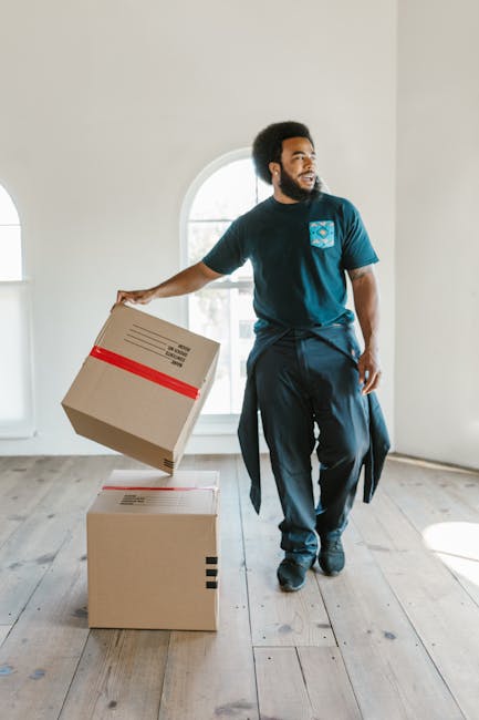 A man with dark skin and an afro hairstyle is inside a bright, empty room with white walls and large arched windows, which allow natural light to illuminate the space. He is dressed in a dark blue t-shirt and navy trousers, with a jacket tied around his waist. The man is in the process of moving, holding a cardboard box with red tape in his right hand, preparing to lift or place it on the floor. The room’s wooden flooring is visible, and the environment appears clean and uncluttered, consistent with a house clearance or home relocation. On the ground in front of him, there is another unopened cardboard box. The overall scene depicts a professional moving or packing process, possibly part of a furniture transport or house removal service offered by Man with Van Tulse Hill, with the focus on careful handling of packed items for successful relocation in Lambeth.