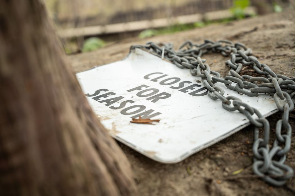 A weathered white sign with bold black lettering reading 'CLOSED FOR SENS...,' partially obscured by a large metal chain draped over it, lies on the ground outdoors. The sign appears dirty and slightly bent, with a small brown leaf resting on its surface. The background shows a wooden fence and soil, with the lower part of a tree trunk visible in the foreground on the left side. The scene is illuminated by natural daylight, indicating an outdoor setting near a property or roadside, where signage related to access restrictions may be relevant to house removals or property management services offered by Man with Van Tulse Hill.