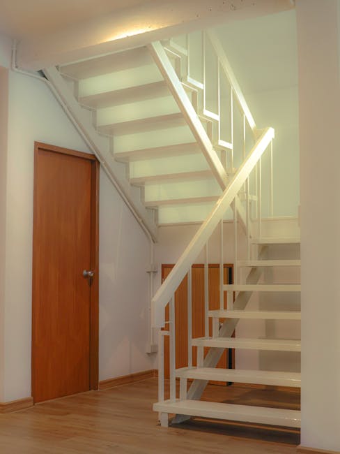 Interior view of a domestic staircase area during a home relocation process, featuring white-painted wooden stairs with open risers, leading to an upper floor. The staircase has a matching white handrail and balustrade, and is positioned adjacent to a light blue wall and a small, square access hatch or vent with a wooden frame and louvers. To the left, there is a closed wooden door with a metallic doorknob, set into a white doorframe, leading to another room. The flooring is light-colored hardwood, consistent throughout the space. There are no objects currently being moved or packed in the visible area, and the lighting appears to be natural or ceiling-mounted, ensuring clear visibility of the staircase and surrounding features. This image relates to house removals and the logistics involved in moving furniture through staircases, as managed by services such as Man with Van Tulse Hill, highlighting the importance of careful planning in stairs, lifts, and size limits during property moves.