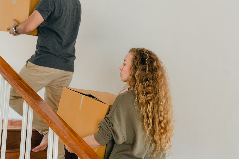 A man with a beard wearing a brown jacket and dark trousers is standing at the top of a wooden staircase inside a house, holding a cardboard box labeled 'Kitchen'. The staircase has a polished wooden handrail and spindles, with a carpeted step visible. Moving blankets are draped over some of the furniture around him, indicating a house removal in progress. A woman dressed in a cream-colored coat and blue jeans is descending the stairs, slightly blurred due to motion. The room behind the man shows a doorway leading to another area and a white ceiling with a smoke detector. Visible in the background are boxed items, packing materials, and part of a vacuum cleaner, all suggesting a packing and moving process. The lighting is natural and bright, highlighting the cluttered yet organized environment typical of house relocation tasks, and aligning with services offered by Man with Van Tulse Hill.