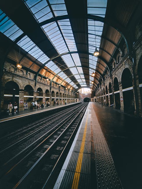 The image depicts the interior of Tulse Hill railway station with a curved, vaulted glass roof overhead, supported by brick and metal arches along both sides of the platform. The platform is lined with tactile paving strips near the edge, and several people are visible sitting or waiting along the side. The railway tracks run through the centre, disappearing into a tunnel at the far end of the station. The station has a historic, industrial appearance with warm lighting illuminating the arched brickwork and the glass roof structure. This setting could be relevant for house removals or moving services involving transportation to or from Tulse Hill, highlighting the logistical aspects of furniture transport and home relocation through railway infrastructure.