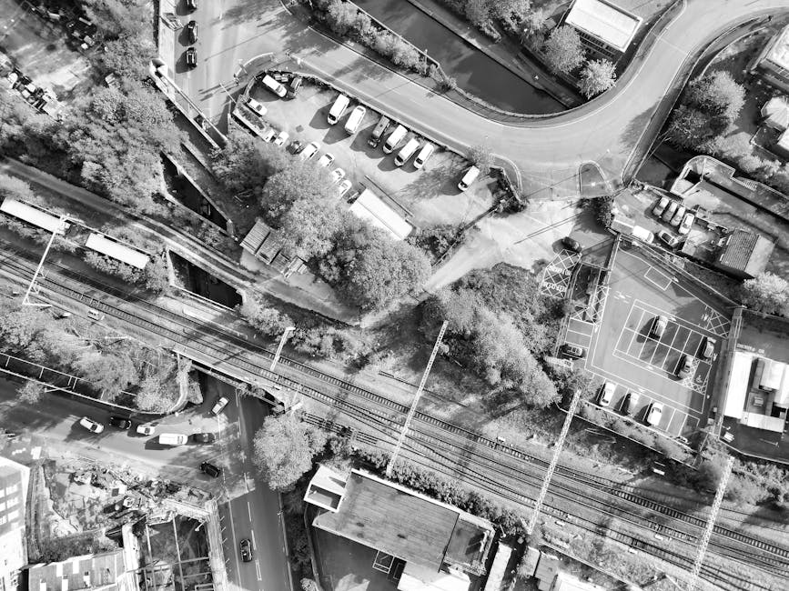 Aerial black-and-white photograph showing a parking lot filled with several parked cars and a playground with a basketball court, surrounded by trees and residential buildings. The parking area is situated near railway tracks, with a narrow path running beside them, and a small structure nearby. In the foreground, a driveway leads into a residential street with additional parked vehicles and houses. The scene captures a typical urban environment, with the railway lines intersecting the area as part of a house relocation or furniture transport process. The setting appears to be in a quiet neighbourhood, with some visible infrastructure supporting moving and loading activities, such as pathways and open spaces near the properties. Man with Van Tulse Hill regularly assists with removals and home relocations in this area, involving careful packing, loading, and transport of furniture and household goods.