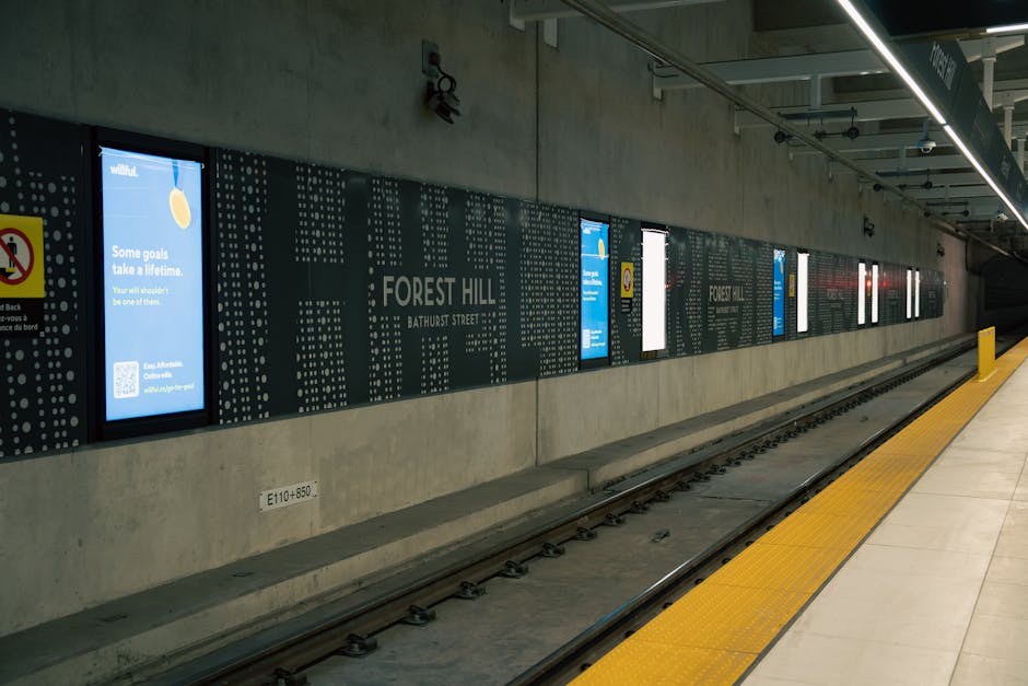 The image depicts the interior of Tulse Hill railway station with a curved, vaulted glass roof overhead, supported by brick and metal arches along both sides of the platform. The platform is lined with tactile paving strips near the edge, and several people are visible sitting or waiting along the side. The railway tracks run through the centre, disappearing into a tunnel at the far end of the station. The station has a historic, industrial appearance with warm lighting illuminating the arched brickwork and the glass roof structure. This setting could be relevant for house removals or moving services involving transportation to or from Tulse Hill, highlighting the logistical aspects of furniture transport and home relocation through railway infrastructure.
