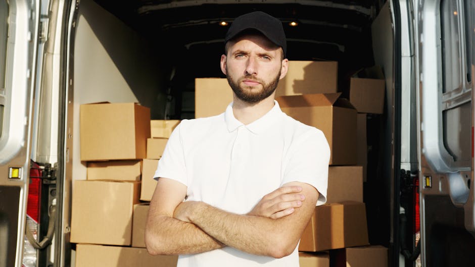 A young man with a beard, wearing a black cap and a white polo shirt, stands inside the open back of a moving van filled with brown cardboard boxes of various sizes, some wrapped in plastic and others exposed, ready for home relocation. The van's interior is dimly lit, with natural light illuminating the man's face and the boxes surrounding him. He has his arms crossed and a serious expression, indicating focus or concern during the packing and loading process. The van is parked outside a residential property, with the background including a paved area and a building partially visible through the open door. The scene captures the packing and furniture transport elements involved in a house removal, aligning with professional moving services such as those offered by Man with Van Tulse Hill, a company specialising in quick and efficient removals and moving logistics.