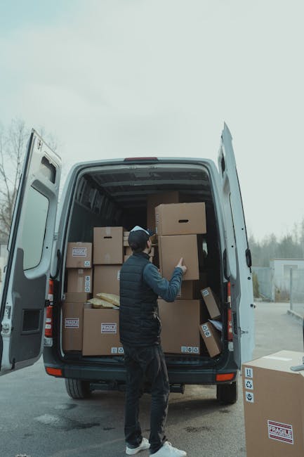A man wearing a dark vest, jeans, and a baseball cap is seen loading or unloading the back of a large white moving van with its rear doors open. The van is parked on a paved area, with several cardboard boxes of varying sizes inside, some stacked on top of each other, and secured with packing materials like bubble wrap or paper. The boxes have labels and barcodes visible, indicating their contents and handling instructions. Nearby, on the ground, there is an additional box. The surroundings include a blurred outdoor background with trees and buildings, suggesting an exterior home or commercial property setting. The scene reflects the process of furniture transport and home relocation, with the individual possibly engaged in packing and moving activities as part of a professional removals service. Man with Van Tulse Hill specializes in house removals and is shown here assisting with an efficient loading process for a quick move, appropriate for urban student relocations or similar household moves.