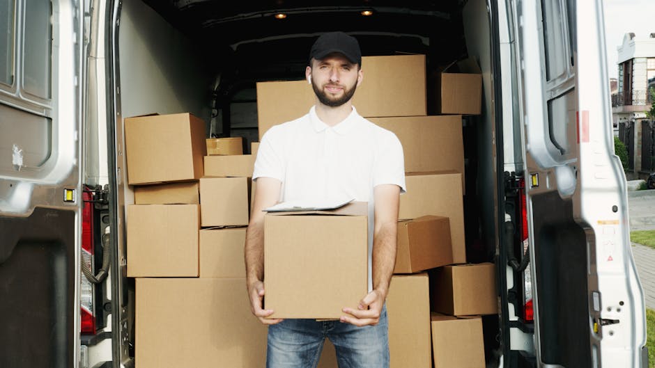 A young man with a beard, wearing a black cap and a white polo shirt, stands inside the open back of a moving van filled with brown cardboard boxes of various sizes, some wrapped in plastic and others exposed, ready for home relocation. The van's interior is dimly lit, with natural light illuminating the man's face and the boxes surrounding him. He has his arms crossed and a serious expression, indicating focus or concern during the packing and loading process. The van is parked outside a residential property, with the background including a paved area and a building partially visible through the open door. The scene captures the packing and furniture transport elements involved in a house removal, aligning with professional moving services such as those offered by Man with Van Tulse Hill, a company specialising in quick and efficient removals and moving logistics.